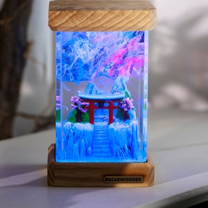 Könnte beinhalten: Rechteckige dekorative Lampe mit japanischem Design. Sie zeigt ein rotes Torii, Berge und eine Treppe in einem transparenten Harzblock. Ober- und Unterseite sind aus Holz. Das Licht strahlt blaue und rosa Farbtöne aus. Der Markenname "BREAKWOODEN" ist sichtbar.