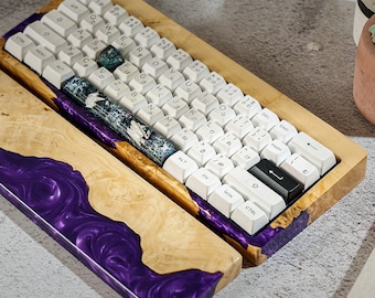 May include: Custom mechanical keyboard with a wooden frame and purple resin accents. The keyboard features white keycaps with Japanese characters and a decorative wrist rest. The design incorporates natural wood grain and a unique, artistic aesthetic.