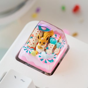 May include: A custom keycap with a cartoon character and decorative elements. The keycap is pink, white, and blue, with a clear top. The keycap is on a white keyboard. The keycap's design includes a cartoon character.