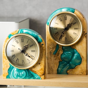 May include: Two decorative mantel clocks with gold-tone faces and Roman numerals. The clocks are set in arched wooden frames with teal resin accents. The wood has a natural grain, and the clocks sit on a light-colored wooden shelf.