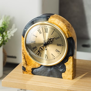 May include: A decorative wooden clock with a gold-colored face and Roman numerals. The clock's frame is a mix of light and dark wood, with a black accent. The clock sits on a wooden surface.