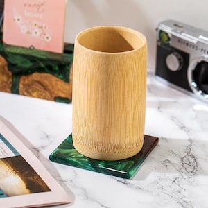 May include: A bamboo cup sits on a square coaster with a green and dark green marbled design. The cup is cylindrical and made of light brown bamboo. The coaster is on a white marble surface.