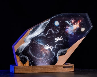 Puede incluir: Una pieza de arte única con temática espacial que presenta un diseño de galaxia con astronautas. La obra de arte está encerrada en una resina transparente y angular, colocada sobre una base de madera. La pieza tiene una luz azul en la base y el texto "BREAKWOODEN".