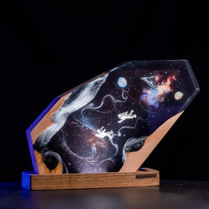 Pode incluir: Uma peça de arte única com tema espacial, apresentando um design de galáxia com astronautas. A obra de arte é envolta em resina transparente e angular, colocada sobre uma base de madeira. A peça tem uma luz azul na base e o texto "BREAKWOODEN".