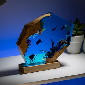 May include: A unique resin art piece depicting an underwater scene with a shipwreck, coral, and marine life. The artwork is encased in a clear, blue resin, with a wooden base and sides. The scene includes a turtle and divers.
