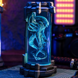 Puede incluir: Una detallada escultura en tonos azules de una criatura Alien encerrada en un recipiente cilíndrico de vidrio. El recipiente está colocado dentro de una base y parte superior oscuras y geométricas, con una luz azul brillante que ilumina la figura alienígena.