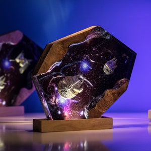 May include: A unique, handcrafted resin lamp featuring a galaxy design with embedded starships. The lamp has a wooden base and frame, with a detailed space scene illuminated by internal lighting. The design includes a Millennium Falcon.