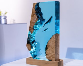 May include: A decorative resin sculpture featuring a marine scene with whales and divers. The sculpture has a light blue center with whales and divers, framed by wood and set on a wooden base. The wood has a natural, light brown color.