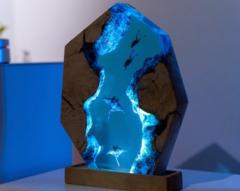 May include: A unique, illuminated resin sculpture featuring a marine scene. The artwork depicts divers and manta rays in a blue, glowing underwater environment, encased within a wooden frame. The base is a rectangular wooden stand.