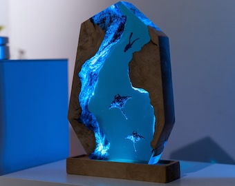 May include: A decorative wooden lamp with a blue, glowing resin interior depicting a marine scene with manta rays and divers. The lamp has a rectangular wooden base and a faceted, geometric shape.