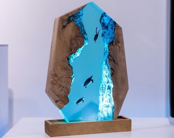 May include: A unique wooden sculpture with a blue resin river design. The sculpture features a light blue resin river with silhouettes of divers and marine life. The wooden frame has a natural wood grain and sits on a wooden base.