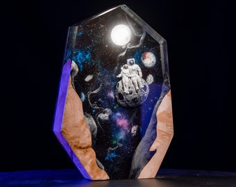 Può includere: Un'opera d'arte in resina unica con una scena spaziale con due astronauti seduti su una luna. L'opera è racchiusa in una forma geometrica trasparente con dettagli in legno. Lo sfondo raffigura una galassia vibrante con pianeti e stelle.
