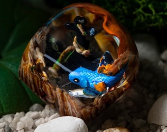 May include: A decorative resin paperweight featuring a blue and orange fish inside a clear, rounded enclosure. The paperweight has a wood-like appearance with a mix of brown, orange, and white tones. The background includes white pebbles and green foliage.