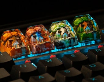 May include: Four custom artisan keycaps featuring miniature house scenes encased in clear resin. Each keycap depicts a different season with vibrant colors: autumn, spring, summer, and winter. The keycaps are installed on a black keyboard with blue backlighting.