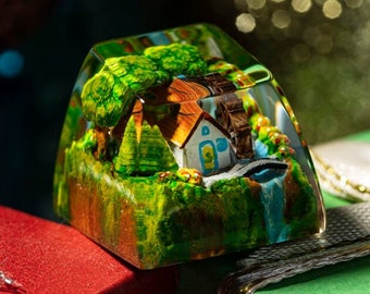 May include: A miniature diorama of a quaint village scene encased in clear resin. The scene includes a small house, trees, a water wheel, and a waterfall. The diorama is set on a green and brown landscape, with a red and green gift box in the background.