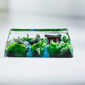 May include: A clear resin keycap featuring a miniature landscape. The scene includes green foliage, gray rocks, a blue waterfall, and a red and black torii gate. The keycap is rectangular with rounded edges.