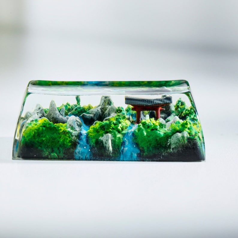 May include: A clear resin keycap featuring a miniature landscape. The scene includes green foliage, gray rocks, a blue waterfall, and a red and black torii gate. The keycap is rectangular with rounded edges.