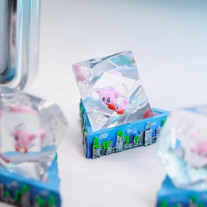 May include: A clear resin sculpture of a pink cartoon character with red gloves, set within a blue box with a cityscape design. The character is suspended within the clear resin, creating a 3D effect.