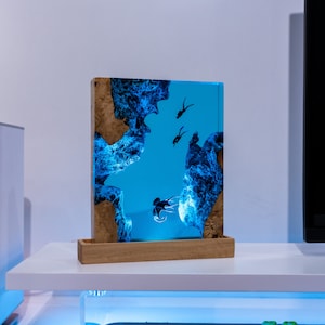 Könnte beinhalten: Ein blaues Kunstharz-Kunstwerk mit einem Holzrahmen. Das Harz zeigt eine blaue Ozeanszene mit Silhouetten von Tauchern und Fischen.