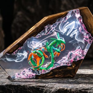 May include: A decorative resin and wood display piece featuring a green serpentine creature and an orange humanoid figure encased in clear resin. The structure includes dark wood edges, pink textured rock-like accents, and swirling white smoke effects.