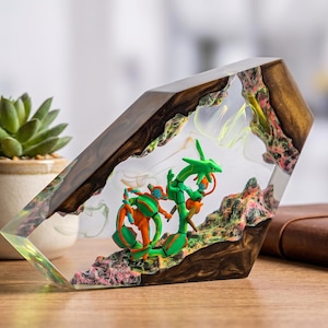 May include: A clear resin block containing a miniature green serpentine creature and an orange and blue humanoid figure. The resin is encased in dark brown wood with textured, multi-colored rocky terrain at the base of the figures.
