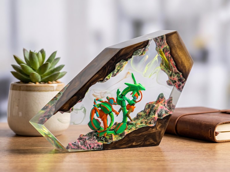 May include: A clear resin block containing a miniature green serpentine creature and an orange and blue humanoid figure. The resin is encased in dark brown wood with textured, multi-colored rocky terrain at the base of the figures.