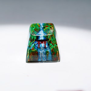 May include: A rectangular keycap with a vibrant landscape design. The keycap features a waterfall scene with a red torii gate, surrounded by lush green and brown foliage. The design is encased in a clear, glossy material.
