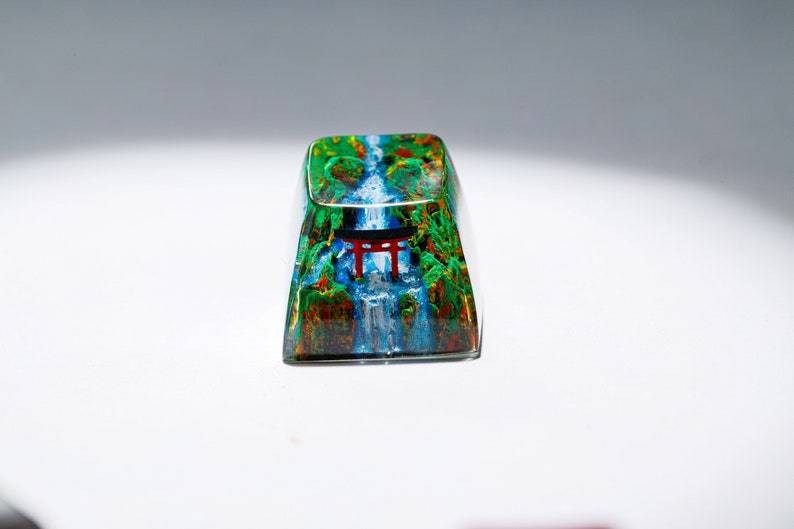 May include: A rectangular keycap with a vibrant landscape design. The keycap features a waterfall scene with a red torii gate, surrounded by lush green and brown foliage. The design is encased in a clear, glossy material.