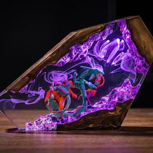 May include: A decorative resin and wood display piece featuring a translucent purple interior with swirling smoke-like patterns. Inside the clear resin, two small, detailed green and orange serpentine figures are positioned among textured purple rock formations.