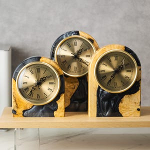 May include: Three decorative clocks with gold-colored faces and black Roman numerals. The clock faces are set in a wooden frame with a unique black and yellow pattern. The clocks are displayed on a wooden shelf.