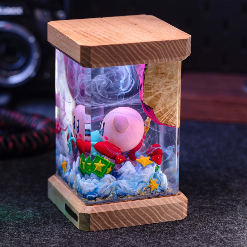 May include: A rectangular resin lamp featuring a pink Kirby character figurine encased in clear resin with swirling white smoke effects. The base and top are made of light-colored wood. The scene includes blue sculpted waves, green blocks with yellow stars, and small yellow star accents.
