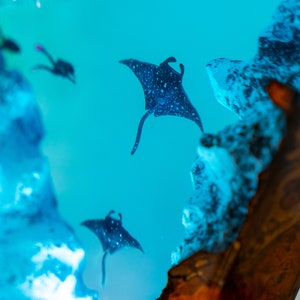 May include: A close-up of a resin art piece with a blue background and a white, cloudy texture.  The resin art piece features three black manta rays swimming in the blue water.