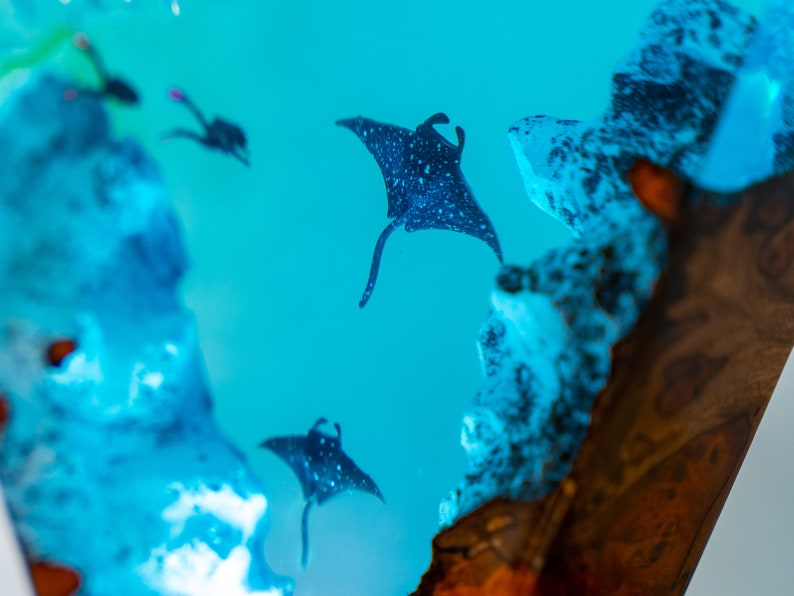 May include: A close-up of a resin art piece with a blue background and a white, cloudy texture.  The resin art piece features three black manta rays swimming in the blue water.