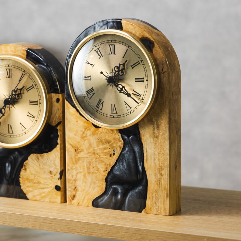 May include: Two wooden mantel clocks with gold-colored clock faces and Roman numerals. The clocks have a natural wood grain finish with black resin accents. The clocks are displayed on a light-colored wooden shelf.