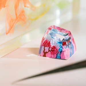 Puede incluir: Una colorida y art&iacute;stica tecla de resina con un dise&ntilde;o de paisaje con un &aacute;rbol floreciente rojo, monta&ntilde;as azules y un cielo rosa y azul. La tecla tiene forma trapezoidal y parece hecha a mano.