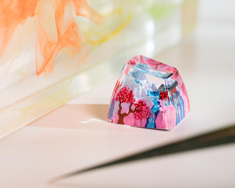Puede incluir: Una colorida y art&iacute;stica tecla de resina con un dise&ntilde;o de paisaje con un &aacute;rbol floreciente rojo, monta&ntilde;as azules y un cielo rosa y azul. La tecla tiene forma trapezoidal y parece hecha a mano.