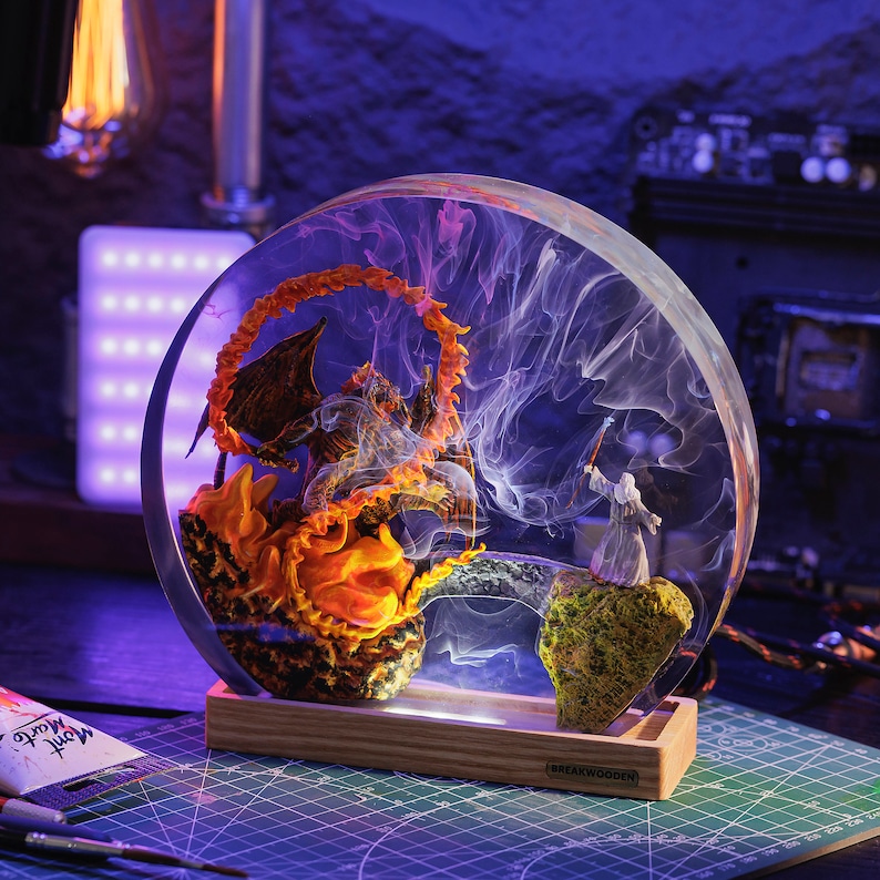May include: A round, clear resin sculpture featuring a fiery demon and a wizard figure. The sculpture is set on a wooden base with a light, creating a dramatic display. The artwork is detailed with vibrant orange, yellow, and white colors.