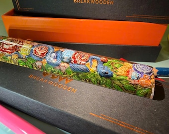 May include: A decorative pencil with a detailed carving of a blue snake, roses, and foliage. The pencil is encased in a clear material, and the design features red, blue, and green colors. The pencil is resting on a black box.