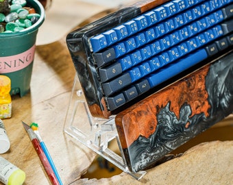 May include: Custom mechanical keyboard with blue and black keycaps, featuring Japanese characters. The keyboard has a unique wooden and resin case with a marbled effect. The keyboard is propped up on a clear acrylic stand.