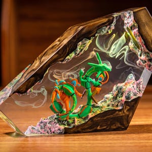 May include: A decorative resin and wood block featuring a green serpentine creature and an orange and blue humanoid figure encased within clear resin. The piece includes textured, rocky elements in pink and green, with wisps of white smoke.