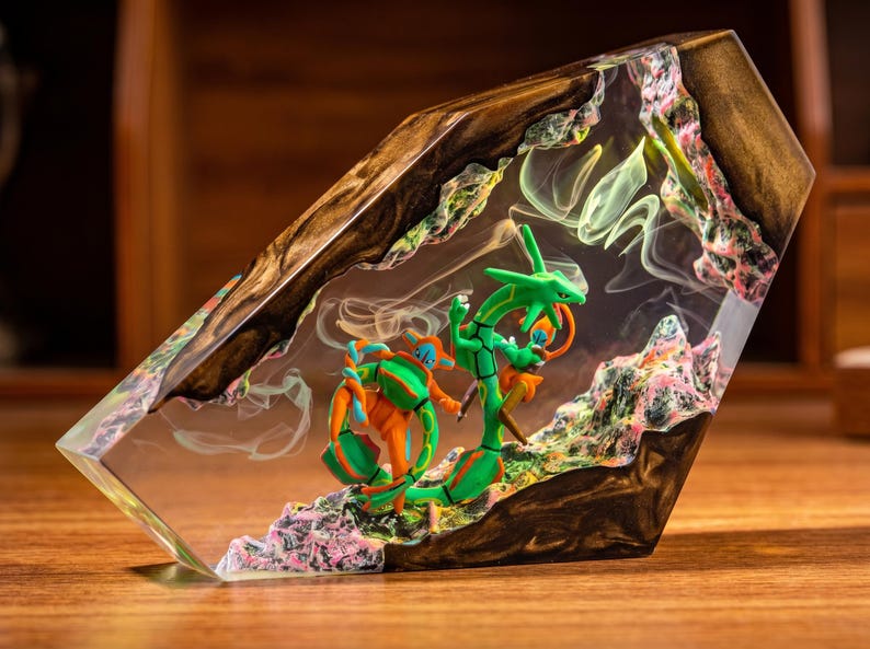 May include: A decorative resin and wood block featuring a green serpentine creature and an orange and blue humanoid figure encased within clear resin. The piece includes textured, rocky elements in pink and green, with wisps of white smoke.