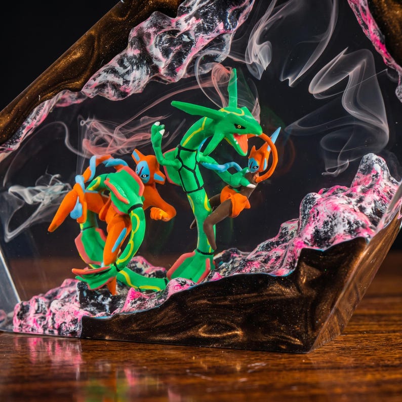 May include: A clear resin diorama featuring a green serpentine creature and an orange humanoid figure with blue accents. The scene is set against a dark, swirling background with pink and black textured rock formations and wisps of white smoke.