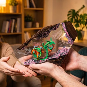 May include: A trapezoidal resin display piece featuring a green serpentine creature and an orange crustacean-like figure embedded within a clear, polished block. The base and sides are textured with dark brown and pink rocky, cavernous details.