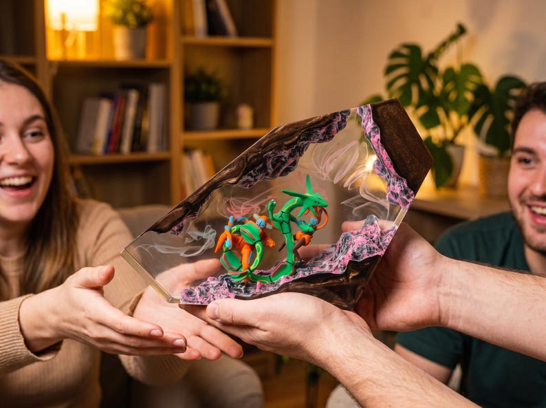 May include: A trapezoidal resin display piece featuring a green serpentine creature and an orange crustacean-like figure embedded within a clear, polished block. The base and sides are textured with dark brown and pink rocky, cavernous details.