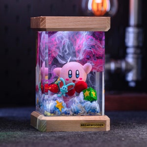 May include: A rectangular resin lamp featuring a pink Kirby character, a small blue creature, and yellow stars encased in clear resin with swirling pink smoke effects. The lamp has a light wood base with a BREAKWOODEN label and a wood top.