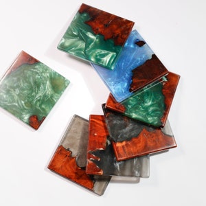 May include: Square coasters with a unique design. Each coaster features a combination of wood and resin in various colors, including green, blue, and gray. The wood has a natural, reddish-brown color, and the resin has a glossy finish.