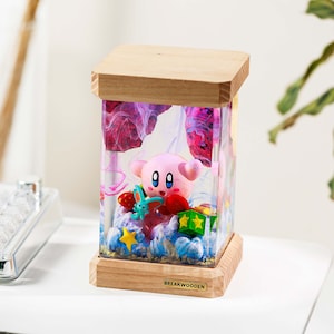 May include: A rectangular resin lamp featuring a pink Kirby figure, a small blue creature, and colorful stars inside. The lamp has a light wood base and top, with the text BREAKWOODEN printed on a small gold plaque on the front wooden base.