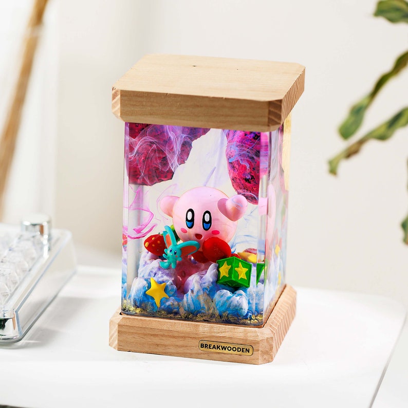May include: A rectangular resin lamp featuring a pink Kirby figure, a small blue creature, and colorful stars inside. The lamp has a light wood base and top, with the text BREAKWOODEN printed on a small gold plaque on the front wooden base.