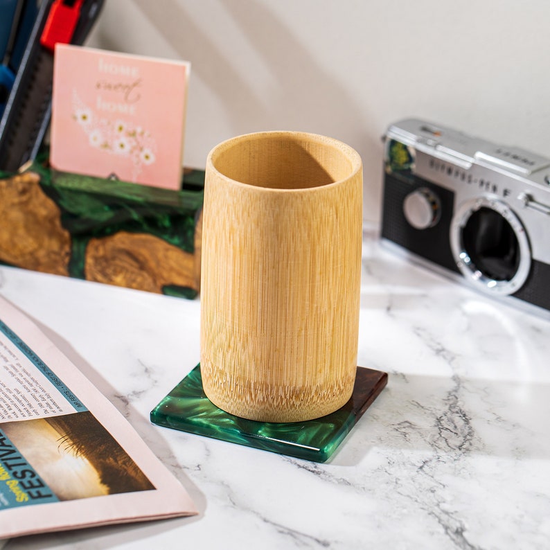 May include: A bamboo cup sits on a square coaster with a green and dark green marbled design. The cup is cylindrical and made of light brown bamboo. The coaster is on a white marble surface.