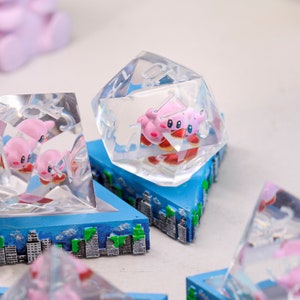 May include: Transparent, multi-faceted dice featuring pink Kirby characters, set on blue bases with cityscape designs. The dice have white numbers. The dice are a unique gaming accessory, displayed on a white surface.